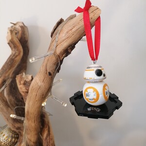 May include: A white and orange Star Wars BB-8 character Christmas bauble with a black base. The bauble is hanging from a red ribbon on a wooden branch.