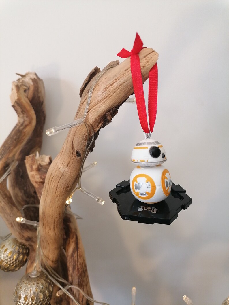 May include: A white and orange Star Wars BB-8 character Christmas bauble with a black base. The bauble is hanging from a red ribbon on a wooden branch.