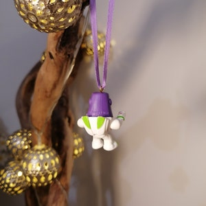 May include: Purple and white toy character ornament hanging from a purple ribbon. The character is a space ranger with a green vest and a laser gun.