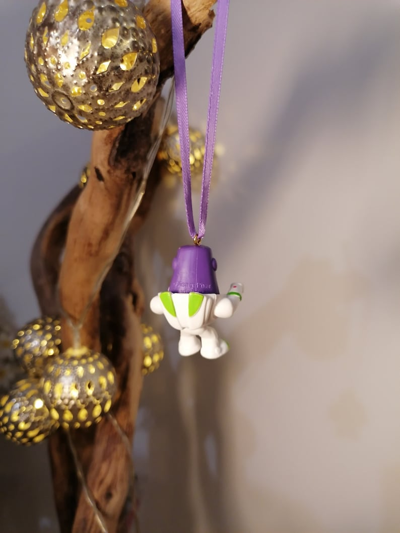 May include: Purple and white toy character ornament hanging from a purple ribbon. The character is a space ranger with a green vest and a laser gun.
