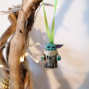 Puede incluir: Un adorno navide&ntilde;o verde y marr&oacute;n con forma de personaje de la franquicia Star Wars. El adorno est&aacute; colgado de una cinta verde.