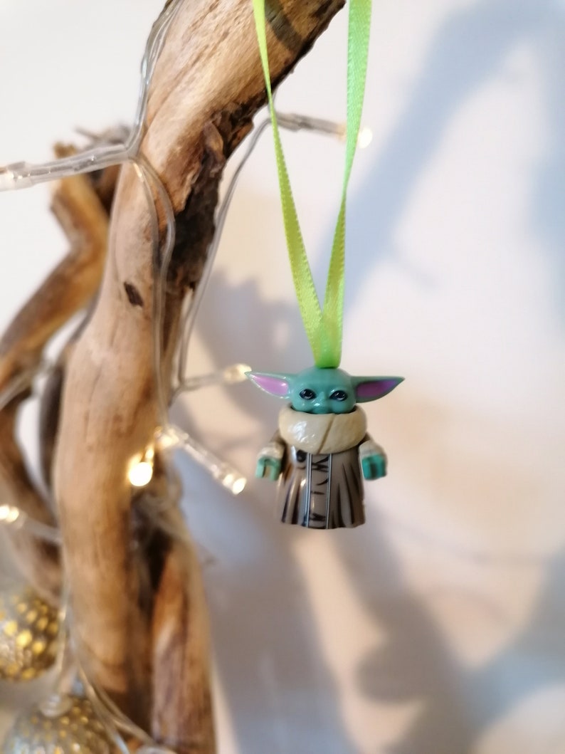 Puede incluir: Un adorno navide&ntilde;o verde y marr&oacute;n con forma de personaje de la franquicia Star Wars. El adorno est&aacute; colgado de una cinta verde.