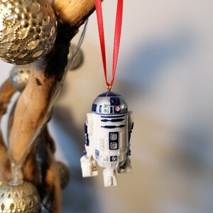K&ouml;nnte beinhalten: Ein wei&szlig;er und blauer R2-D2 Weihnachtsbaumschmuck mit einem roten Band, das an einem Ast h&auml;ngt.