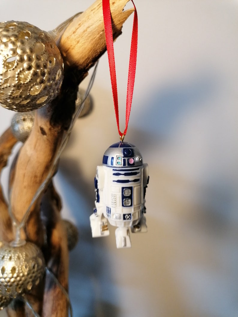 K&ouml;nnte beinhalten: Ein wei&szlig;er und blauer R2-D2 Weihnachtsbaumschmuck mit einem roten Band, das an einem Ast h&auml;ngt.