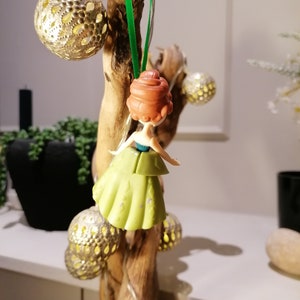 May include: A green and gold Christmas ornament featuring a small plastic doll with red hair wearing a green dress. The ornament is hanging from a green ribbon on a branch.