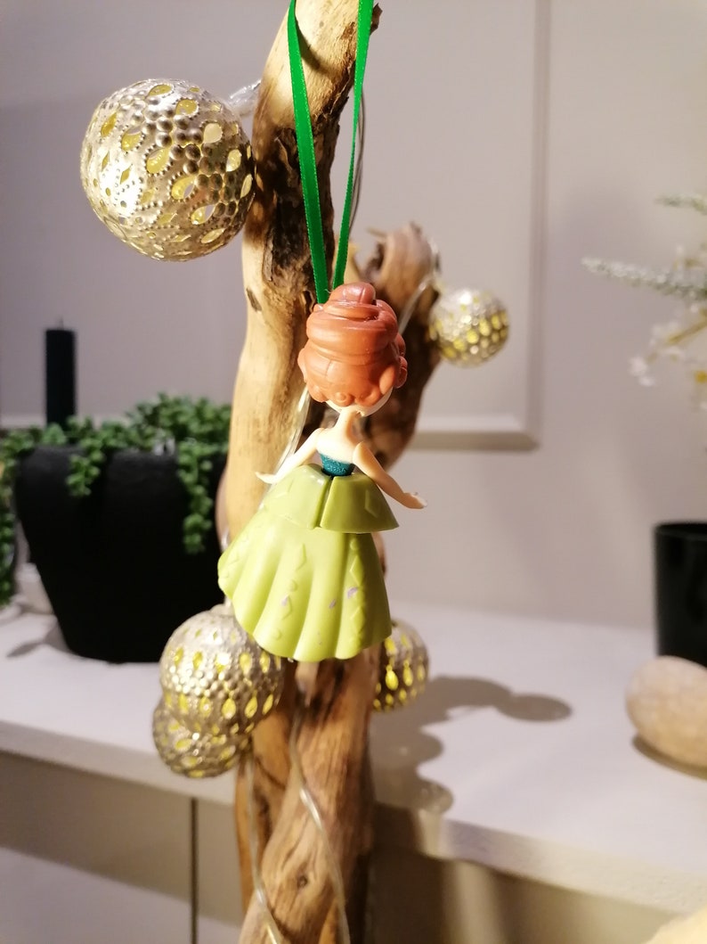 May include: A green and gold Christmas ornament featuring a small plastic doll with red hair wearing a green dress. The ornament is hanging from a green ribbon on a branch.