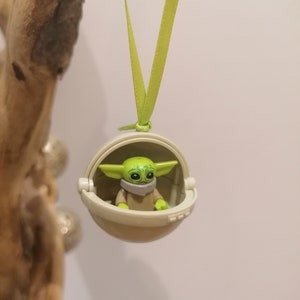May include: A green and tan Baby Yoda figure inside a white pod, hanging from a light green ribbon. The figure has large eyes and pointed ears. The pod has a hinged opening. The background is a light color.