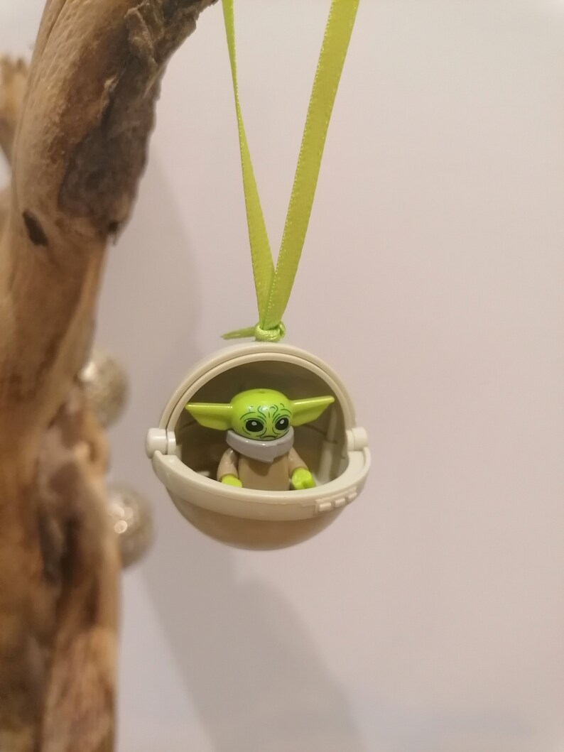 May include: A green and tan Baby Yoda figure inside a white pod, hanging from a light green ribbon. The figure has large eyes and pointed ears. The pod has a hinged opening. The background is a light color.