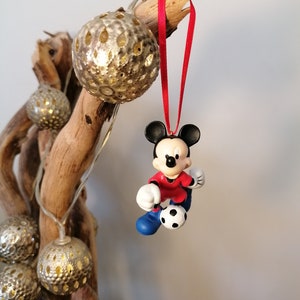 K&ouml;nnte beinhalten: Eine rote und blaue Mickey Mouse Figur, die ein rotes Trikot und blaue Shorts tr&auml;gt, tritt einen Fu&szlig;ball. Die Figur h&auml;ngt an einem roten Band an einem Ast mit goldenen Ornamenten.