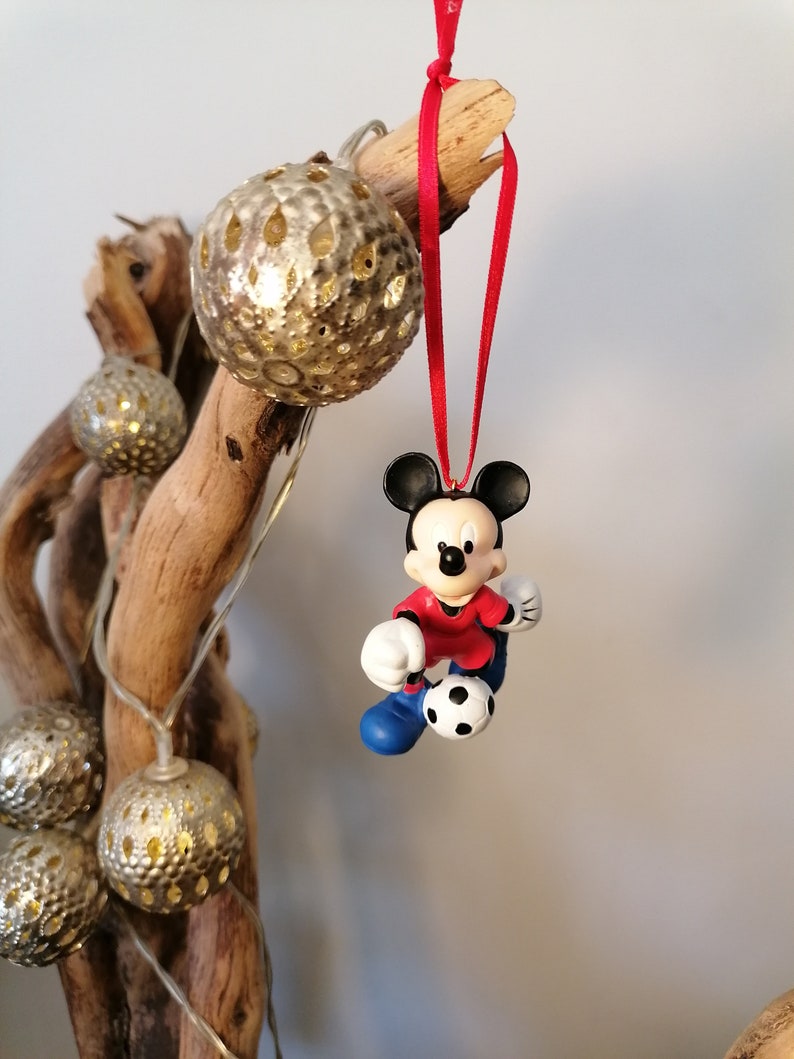 K&ouml;nnte beinhalten: Eine rote und blaue Mickey Mouse Figur, die ein rotes Trikot und blaue Shorts tr&auml;gt, tritt einen Fu&szlig;ball. Die Figur h&auml;ngt an einem roten Band an einem Ast mit goldenen Ornamenten.
