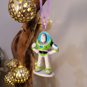 Puede incluir: Una cinta morada cuelga de una rama con un juguete de pl&aacute;stico de Buzz Lightyear de la pel&iacute;cula Toy Story. El juguete es blanco con detalles verdes, azules y amarillos. El juguete est&aacute; colgado de una rama con una cadena de luces.