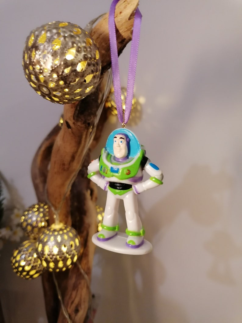 Puede incluir: Una cinta morada cuelga de una rama con un juguete de pl&aacute;stico de Buzz Lightyear de la pel&iacute;cula Toy Story. El juguete es blanco con detalles verdes, azules y amarillos. El juguete est&aacute; colgado de una rama con una cadena de luces.