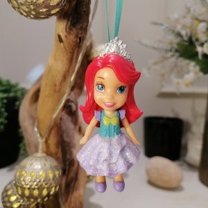 Peut inclure: Une poup&eacute;e princesse aux cheveux roux avec une robe violette et une couronne argent&eacute;e, suspendue &agrave; un ruban turquoise.