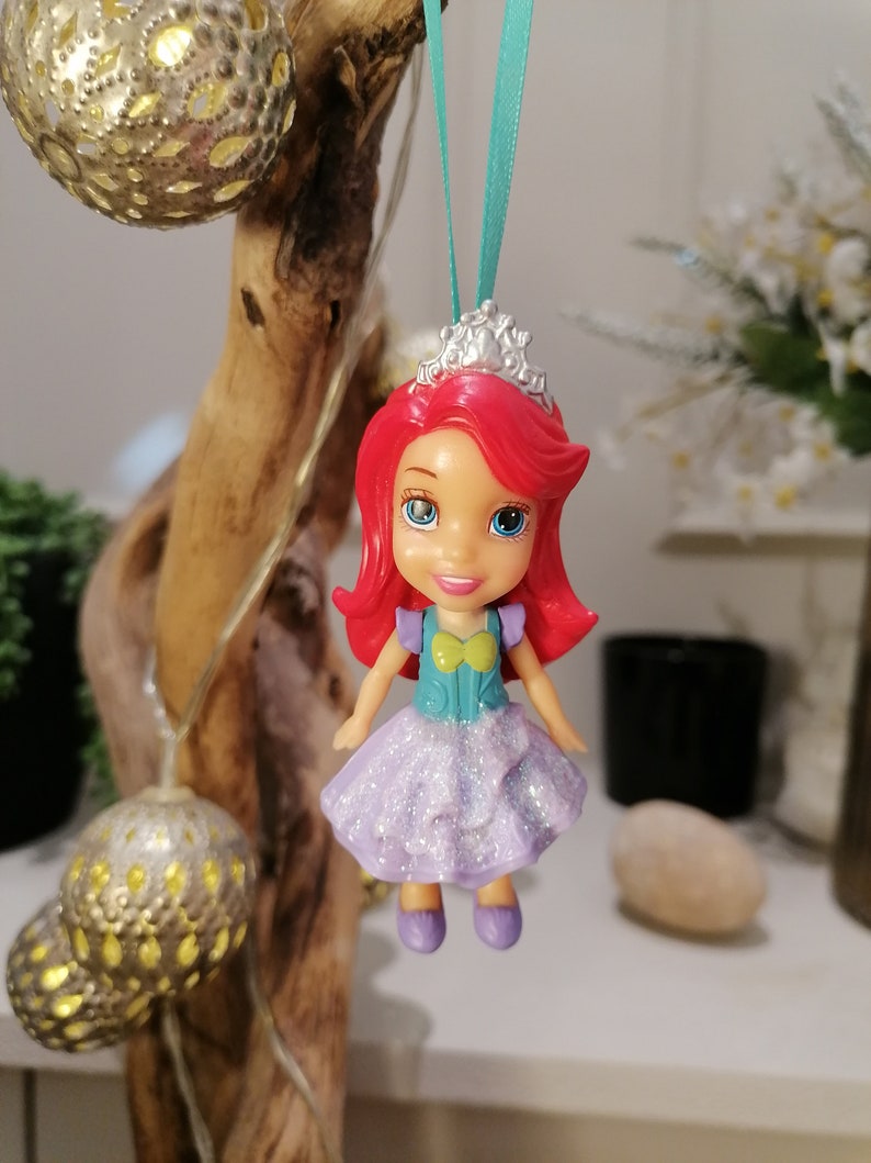Peut inclure: Une poup&eacute;e princesse aux cheveux roux avec une robe violette et une couronne argent&eacute;e, suspendue &agrave; un ruban turquoise.