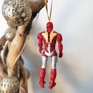 Pode incluir: Enfeite de Natal vermelho e prateado com uma figura de super-her&oacute;i e uma fita dourada.