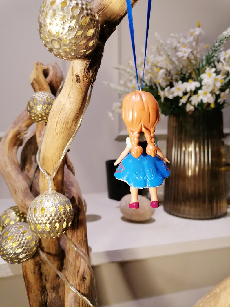 K&ouml;nnte beinhalten: Eine kleine Plastikpuppe mit roten Haaren in Z&ouml;pfen, die ein blaues Kleid mit wei&szlig;en Akzenten tr&auml;gt, das an einem blauen Band h&auml;ngt. Die Puppe h&auml;ngt an einem Ast mit goldenen, metallischen Ornamenten.