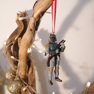 May include: A green and gray Star Wars Boba Fett action figure ornament hanging from a red ribbon on a branch with white lights.