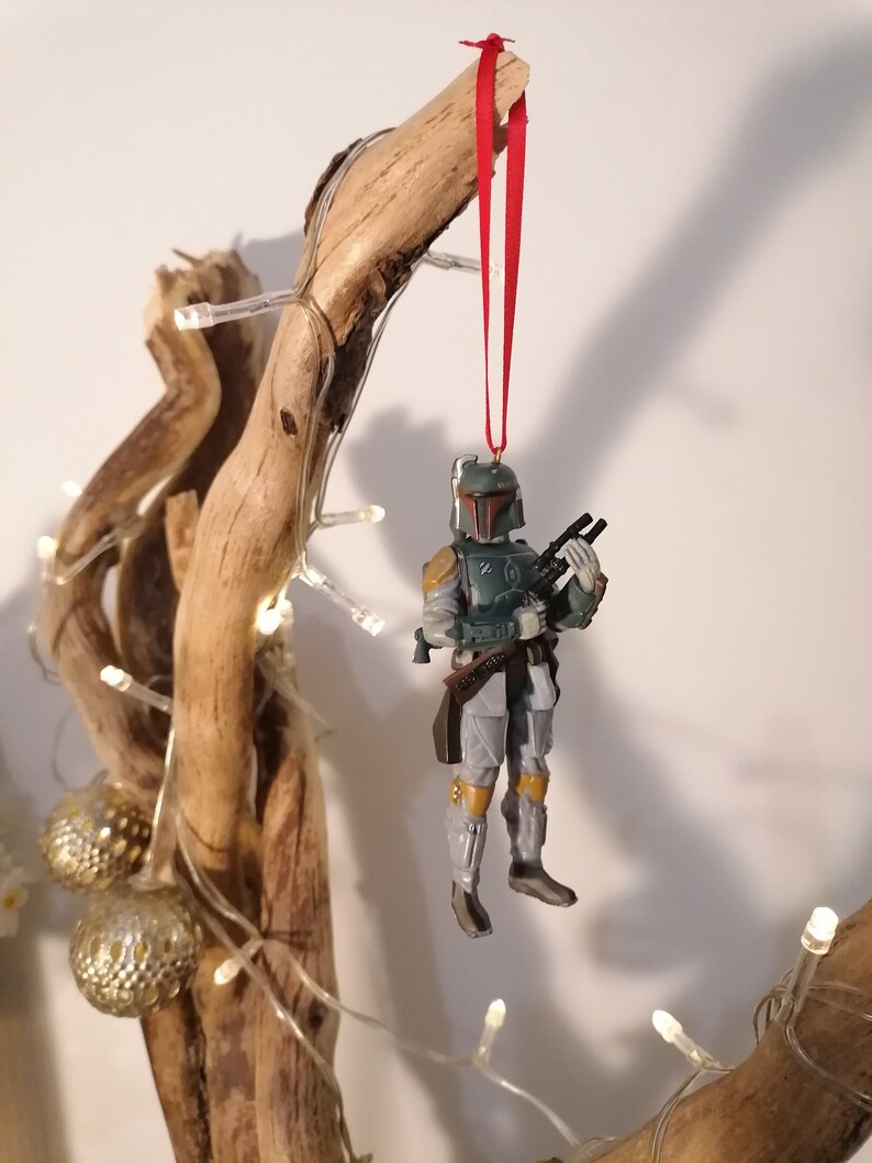 May include: A green and gray Star Wars Boba Fett action figure ornament hanging from a red ribbon on a branch with white lights.