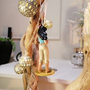 May include: A figurine of a woman with long black hair wearing a blue tank top and white pants. She is standing on a yellow base and is hanging from a white string.