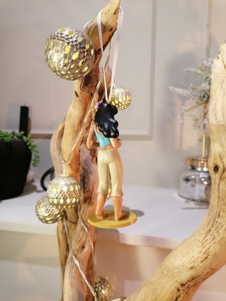 May include: A figurine of a woman with long black hair wearing a blue tank top and white pants. She is standing on a yellow base and is hanging from a white string.