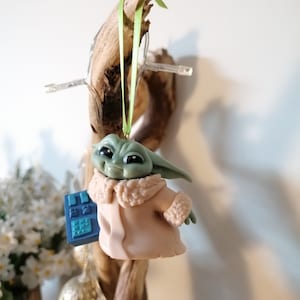 May include: A green and brown figurine of a character from the Star Wars franchise, wearing a tan coat and holding a blue device. The figurine is hanging from a branch with a green ribbon.