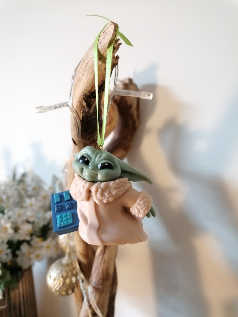 May include: A green and brown figurine of a character from the Star Wars franchise, wearing a tan coat and holding a blue device. The figurine is hanging from a branch with a green ribbon.