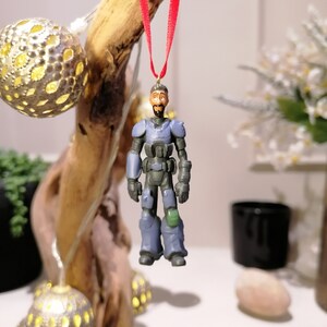 Puede incluir: Un adorno de figura de acción azul y verde de un hombre con casco y sosteniendo un arma. El adorno está colgado de una cinta roja.