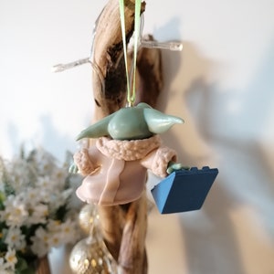 May include: A green and brown Yoda ornament wearing a pink robe and holding a blue Lego brick. The ornament is hanging from a branch.