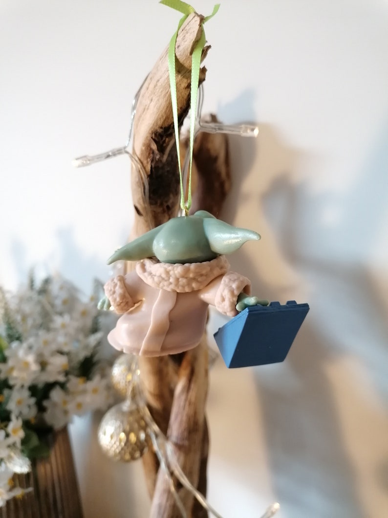 May include: A green and brown Yoda ornament wearing a pink robe and holding a blue Lego brick. The ornament is hanging from a branch.