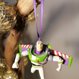 May include: Purple and white Christmas ornament featuring a plastic figurine of Buzz Lightyear from the Toy Story movie franchise. Buzz Lightyear is wearing his signature space ranger suit and has his wings extended.