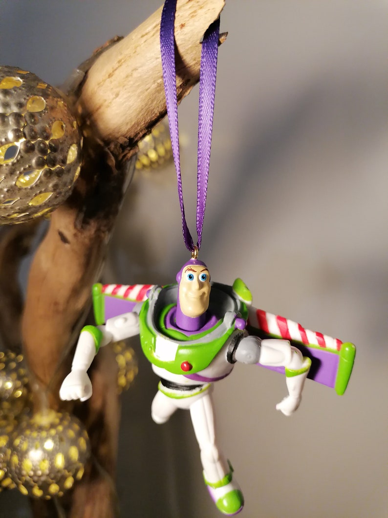 May include: Purple and white Christmas ornament featuring a plastic figurine of Buzz Lightyear from the Toy Story movie franchise. Buzz Lightyear is wearing his signature space ranger suit and has his wings extended.