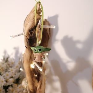 May include: A green and tan Baby Yoda figurine Christmas ornament hanging from a piece of driftwood. The ornament is suspended by a green ribbon. A string of white lights and decorative gold ornaments are also visible.