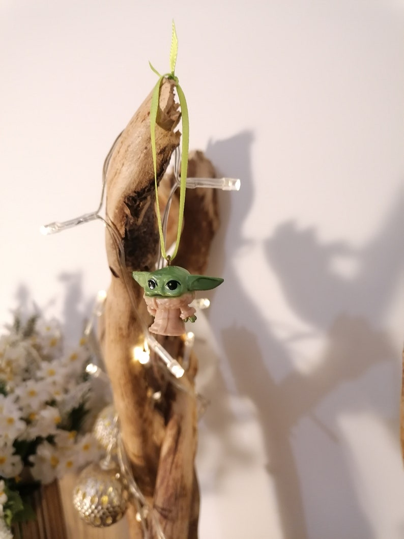 May include: A green and tan Baby Yoda figurine Christmas ornament hanging from a piece of driftwood. The ornament is suspended by a green ribbon. A string of white lights and decorative gold ornaments are also visible.