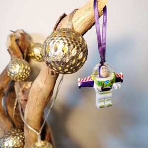May include: A purple ribbon hangs from a branch with a white plastic toy figure of Buzz Lightyear from the Toy Story movie franchise. The figure is wearing a white spacesuit with a purple and red stripe across the chest. The figure is holding a laser gun. The branch is decorated with gold metallic fairy lights.