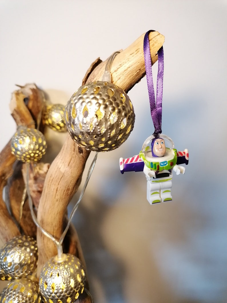 May include: A purple ribbon hangs from a branch with a white plastic toy figure of Buzz Lightyear from the Toy Story movie franchise. The figure is wearing a white spacesuit with a purple and red stripe across the chest. The figure is holding a laser gun. The branch is decorated with gold metallic fairy lights.