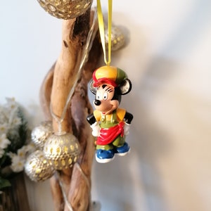 May include: A ceramic figurine of Mickey Mouse wearing a yellow and red hat and a green and red outfit. The figurine is hanging from a yellow ribbon on a branch.