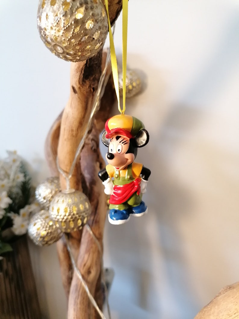 May include: A ceramic figurine of Mickey Mouse wearing a yellow and red hat and a green and red outfit. The figurine is hanging from a yellow ribbon on a branch.