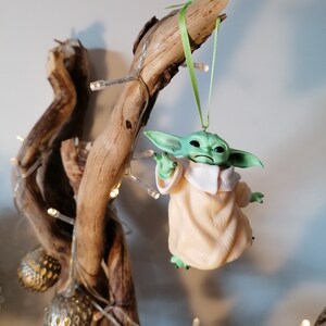 May include: A green and beige figurine of a character from the Star Wars franchise hanging from a branch with a green ribbon. The figurine is hanging from a branch with a string of lights.