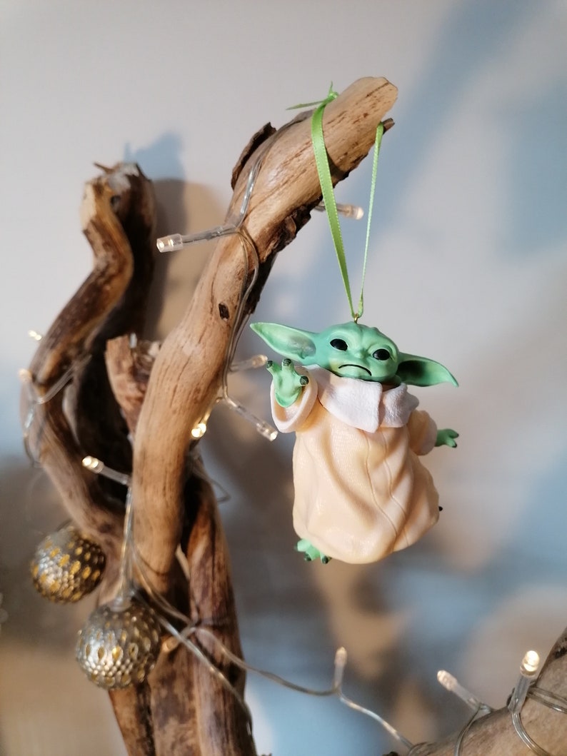 May include: A green and beige figurine of a character from the Star Wars franchise hanging from a branch with a green ribbon. The figurine is hanging from a branch with a string of lights.