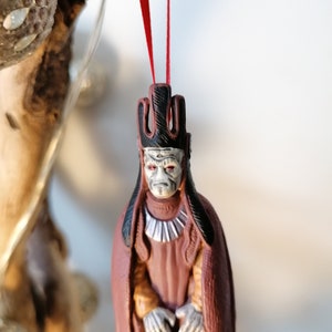 May include: A red and black Christmas ornament featuring a character from Star Wars. The character is wearing a red robe and a black crown. The ornament is hanging from a red ribbon.