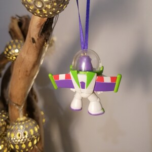 Puede incluir: Un adorno de Buzz Lightyear, ranger espacial, de color morado y verde, con una c&uacute;pula de pl&aacute;stico transparente. El adorno est&aacute; colgado de una cinta morada.