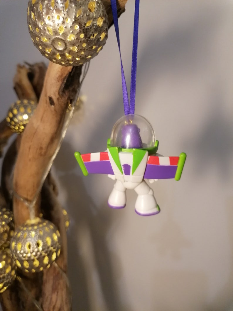 Puede incluir: Un adorno de Buzz Lightyear, ranger espacial, de color morado y verde, con una c&uacute;pula de pl&aacute;stico transparente. El adorno est&aacute; colgado de una cinta morada.