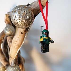 May include: A green and yellow Lego minifigure of Boba Fett hanging from a red ribbon on a driftwood branch with silver Christmas ornaments.