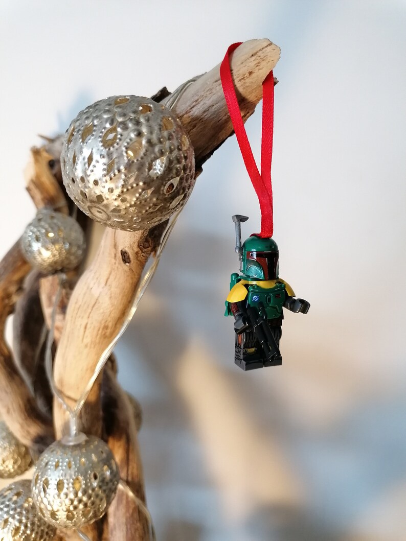 May include: A green and yellow Lego minifigure of Boba Fett hanging from a red ribbon on a driftwood branch with silver Christmas ornaments.