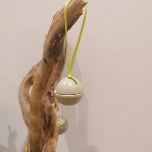 May include: A light gray, spherical ornament suspended by a bright yellow ribbon. The ornament is hanging from a piece of driftwood. Other silver ornaments are visible in the background.