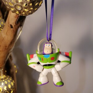 Puede incluir: Un adorno de juguete de pl&aacute;stico morado y verde de Buzz Lightyear de la franquicia de pel&iacute;culas Toy Story. El adorno est&aacute; colgado de una cinta morada.