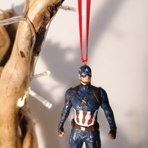 May include: A red ribbon hangs from a branch with a Captain America figurine ornament attached. The figurine is wearing a blue and red costume with a white star on the chest and a red, white, and blue shield on his arm.