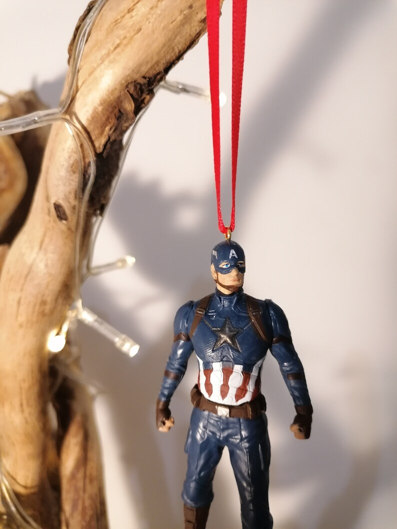 May include: A red ribbon hangs from a branch with a Captain America figurine ornament attached. The figurine is wearing a blue and red costume with a white star on the chest and a red, white, and blue shield on his arm.
