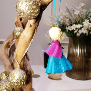 K&ouml;nnte beinhalten: Ein blauer und lilafarbener Plastik-Anh&auml;nger einer Prinzessin mit blonden Haaren. Der Anh&auml;nger h&auml;ngt an einem blauen Band an einem Ast.