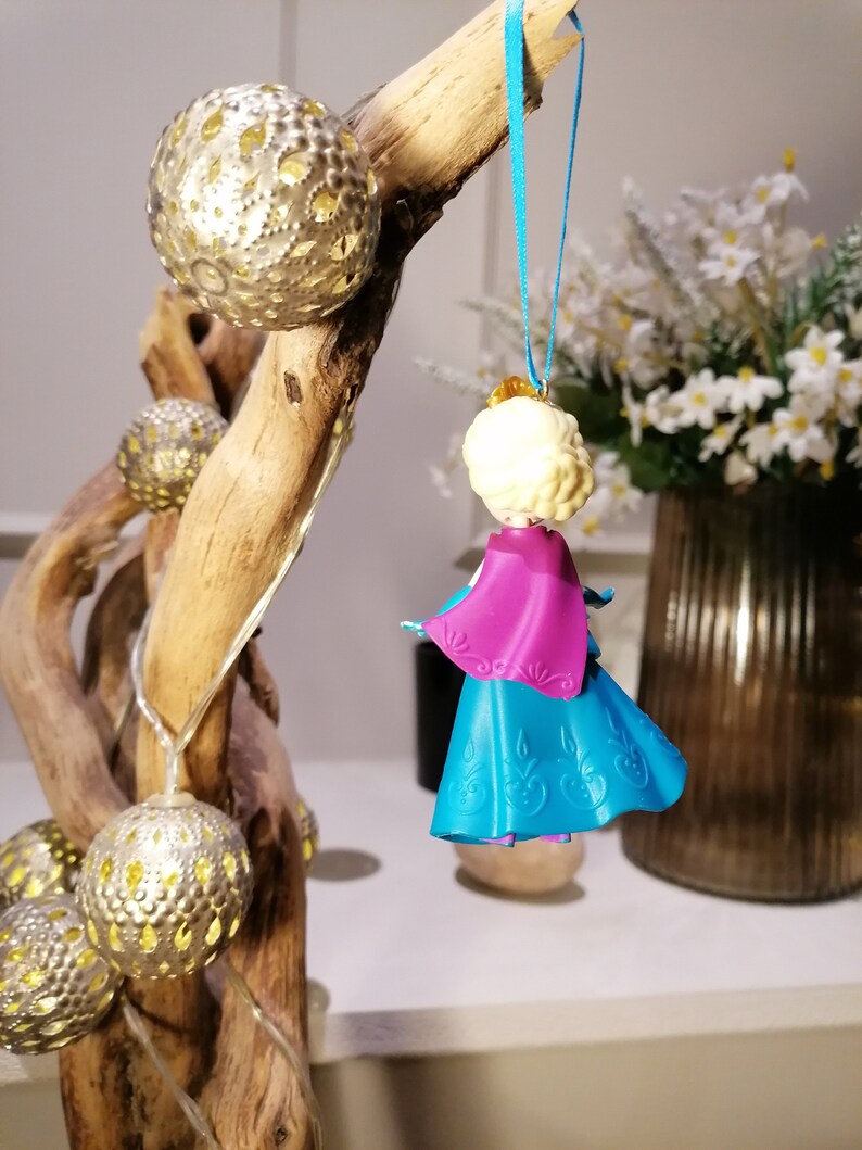 K&ouml;nnte beinhalten: Ein blauer und lilafarbener Plastik-Anh&auml;nger einer Prinzessin mit blonden Haaren. Der Anh&auml;nger h&auml;ngt an einem blauen Band an einem Ast.
